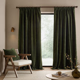 Heath washed velvet pencil pleat curtains in olive green hanging full-length by a window, shown in a living room with a wooden armchair and round coffee table.