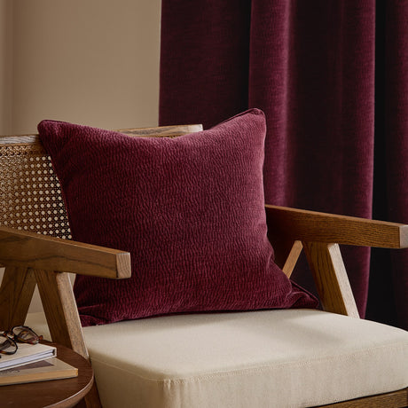 Heath Washed Velvet Jacquard Polyester Cushion in beetroot red, textured square design on a wooden armchair with cane detailing and burgundy curtains behind.