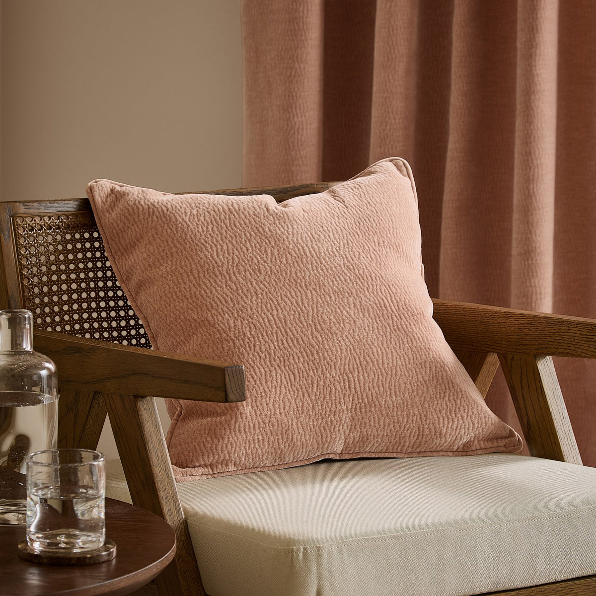 Heath washed velvet jacquard cushion in dusky pink Cotta with piped edge, shown on a wooden armchair beside pink curtains.