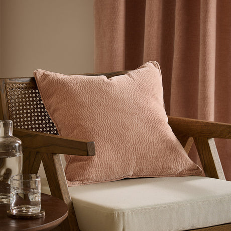 Heath washed velvet jacquard cushion in dusky pink Cotta with piped edge, shown on a wooden armchair beside pink curtains.