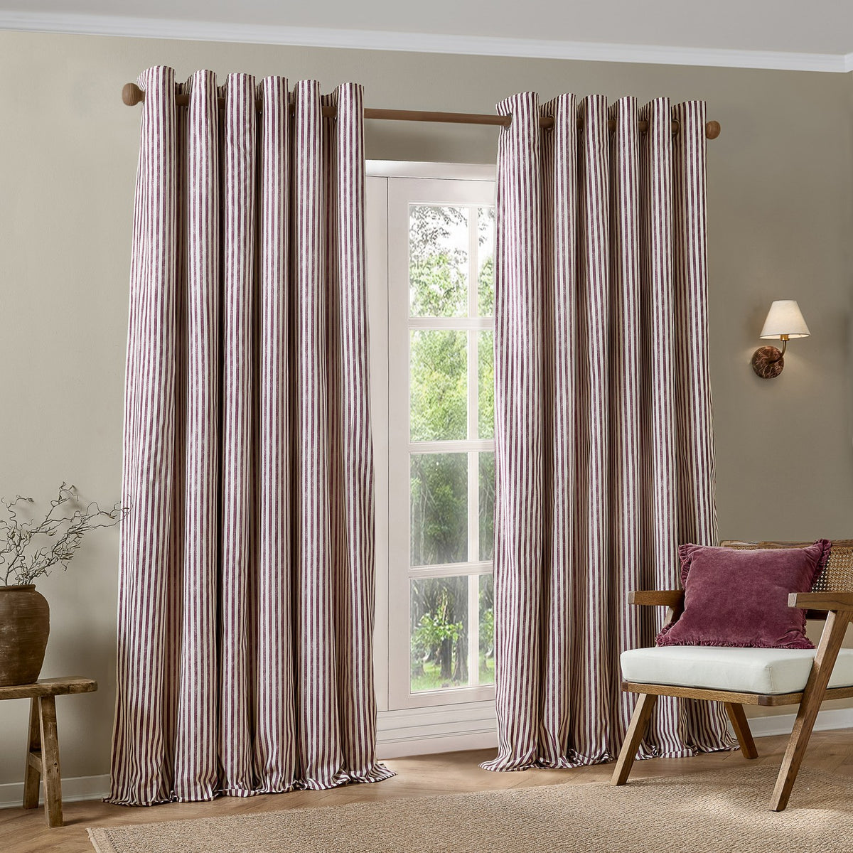 Hebden Cotton Fully Lined Eyelet Curtains in Beetroot, a pair of cream and red vertical striped panels with wooden eyelet rings, shown hung on a wooden pole beside a window in a living room.