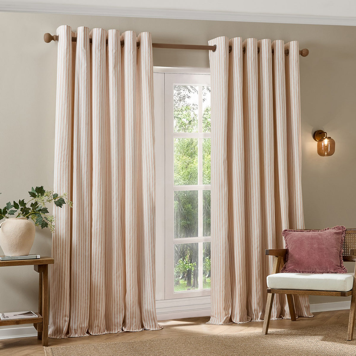 Hebden Cotton Fully Lined Eyelet Curtains in Blush, showing pink and cream striped panels with metal eyelet headers hung on a wooden pole in a living room setting.