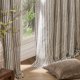 Hebden Cotton Fully Lined Eyelet Curtains in Chocolate, showing brown and cream vertical striped fabric hanging by a window with a fringed cushion nearby.