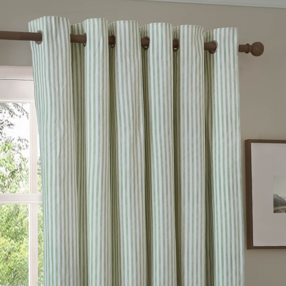 Hebden Cotton Fully Lined Eyelet Curtain in Eucalyptus, green and cream vertical stripe fabric with eyelet heading on a wooden pole, shown in a living room setting.