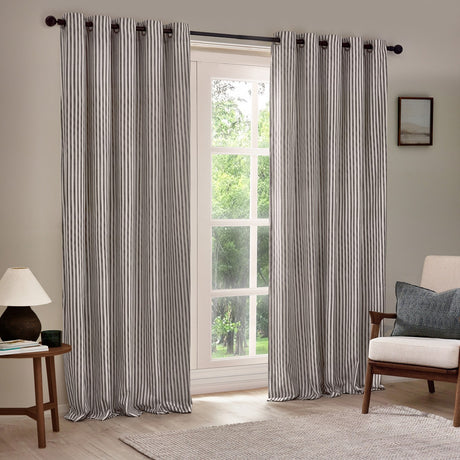 Hebden Cotton Lined Eyelet Curtains in grey, showing narrow vertical stripe fabric with metal eyelet heading, hung on a black pole beside a living room window.