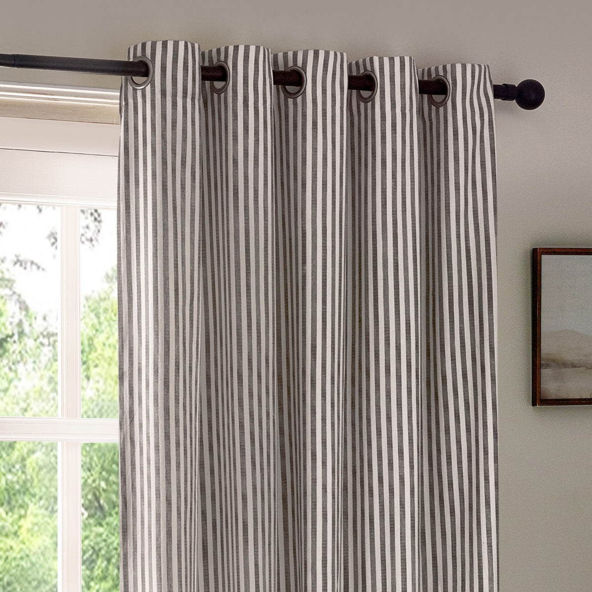 Hebden Cotton Lined Eyelet Curtains in grey, striped panel with metal eyelet heading on a dark pole, hung at a living room window beside a wall-mounted frame.
