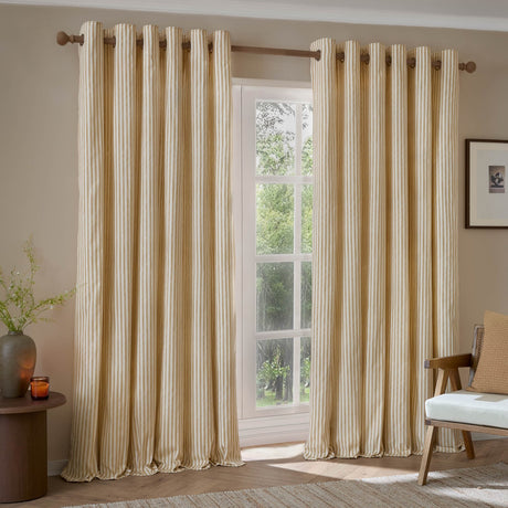 Hebden Cotton Fully Lined Eyelet Curtains in honey yellow stripe on off-white, hung on a wooden pole beside a window in a living room setting.