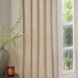 Hebden Cotton Fully Lined Eyelet Curtains in Honey Yellow, vertical striped panels hung by a window beside a vase of foliage in a living room setting.