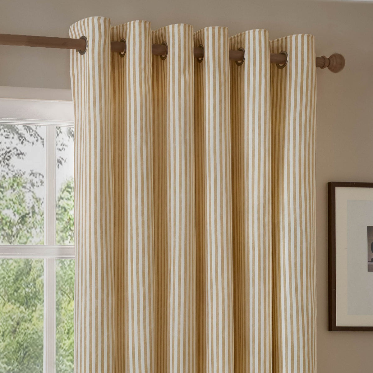 Hebden Cotton Fully Lined Eyelet Curtains in Honey Yellow, vertical striped fabric on a wooden pole with eyelet header, hung beside a window in a living room.