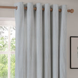 Hebden Cotton Fully Lined Eyelet Curtains in Mineral Blue, showing vertical stripe fabric with metal grommet eyelets threaded onto a wooden pole beside a window in a light-toned room.