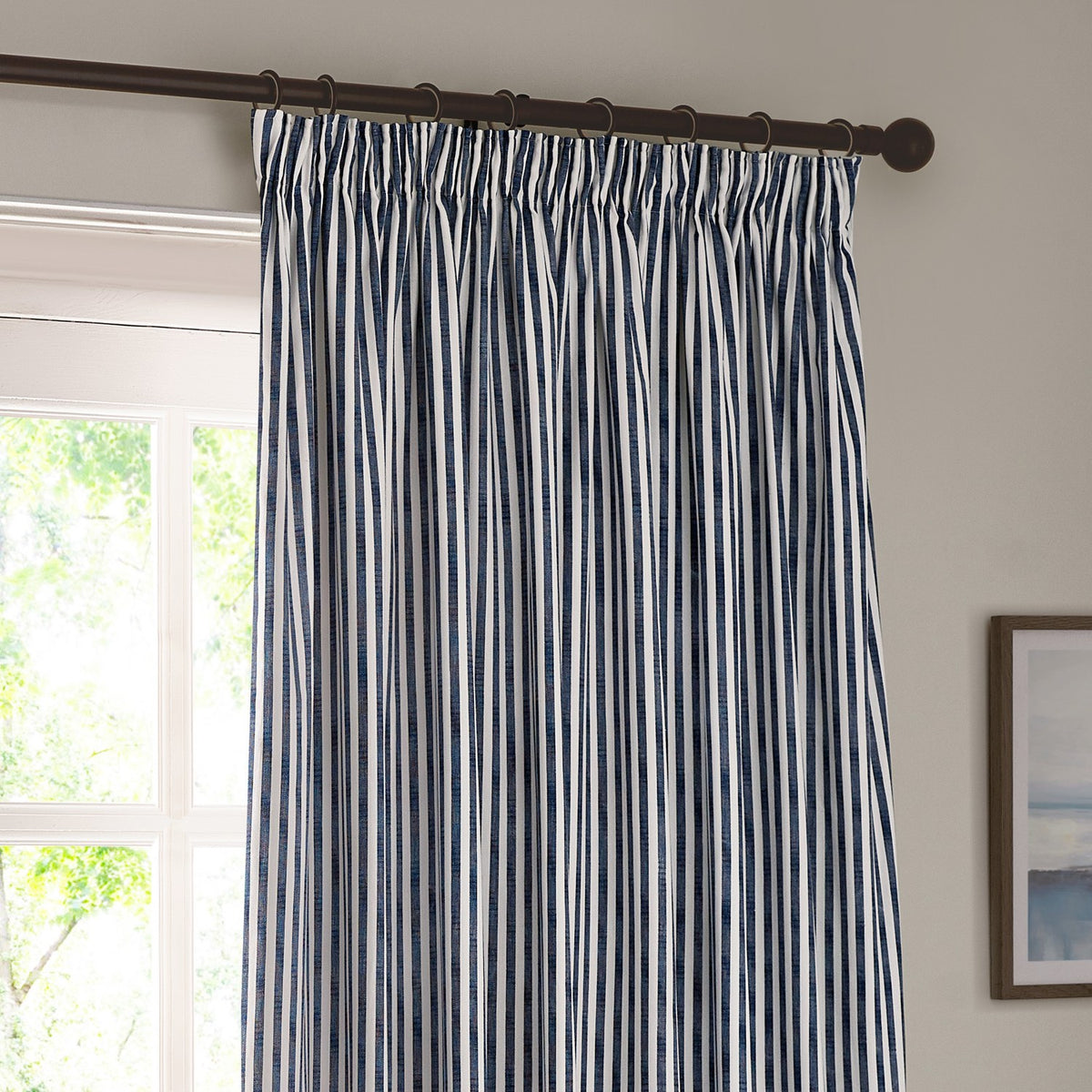 Hebden Cotton Lined Eyelet Curtains in navy and white vertical stripe, shown pencil-pleated on a dark rod in a living room beside a window.