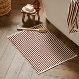 Hebden Striped Cotton Bath Mat in pecan brown and cream, shown on wood flooring beside a bath with a wooden stool, soap, and body brush.