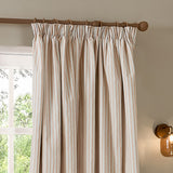 Hebden fully lined cotton pencil pleat curtains in blush and cream stripe, shown gathered on a wooden pole beside a window in a living room setting.
