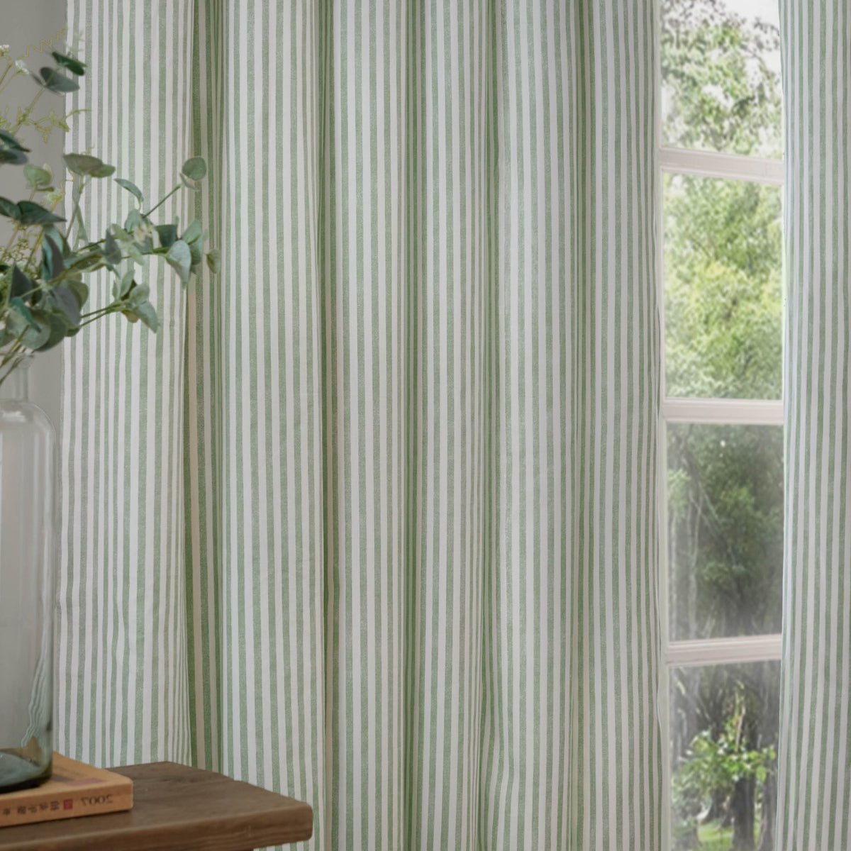 Hebden Fully Lined Cotton Pencil Pleat Curtains in Eucalyptus, showing a green and white striped pair hung at a window beside a plant and wooden table.