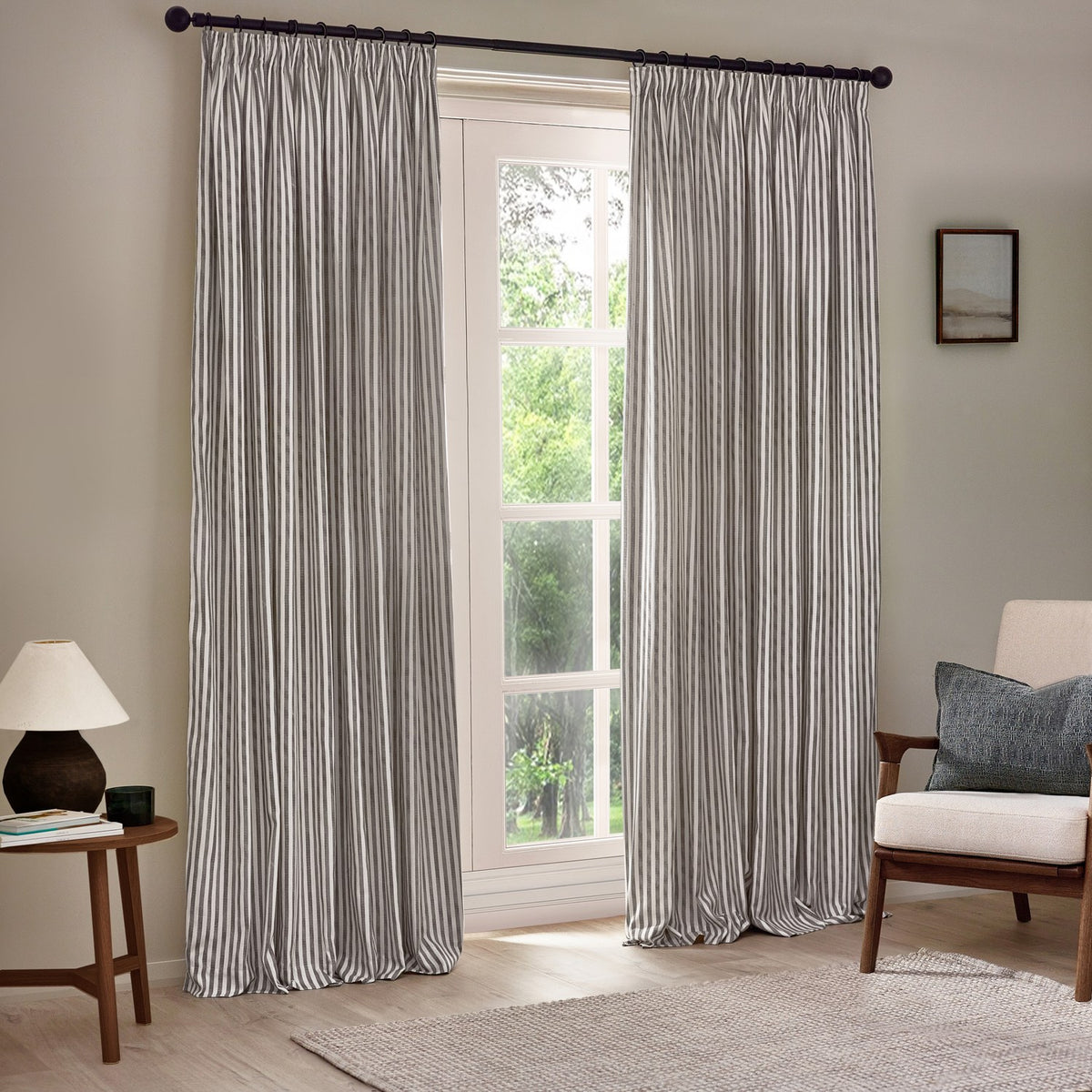 Hebden Striped Cotton Pencil Pleat Curtains in grey and white, hung on a black pole beside a window in a living room with an armchair and side table.