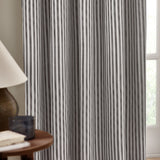 Hebden pencil pleat curtains in grey-and-white striped cotton, hung by a window in a living room with a side table and lamp nearby.
