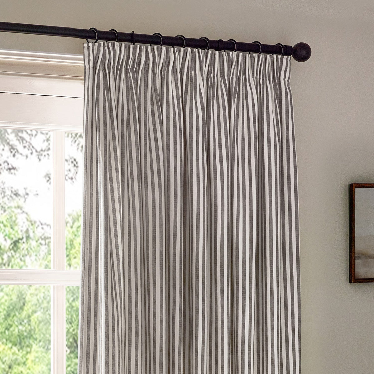 Hebden Striped Cotton Pencil Pleat Curtains in grey and white, hung on a black rod with pinch pleat heading beside a window in a living room.