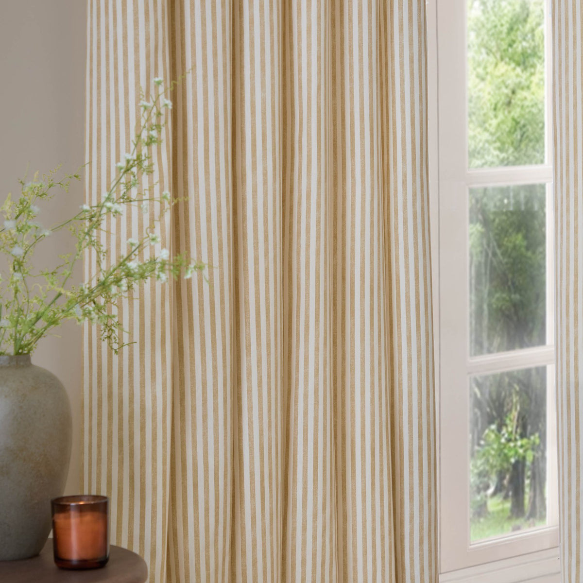 Hebden fully lined pencil pleat curtains in honey yellow and cream vertical stripe cotton, hung beside a window in a living room with a vase and candle nearby.