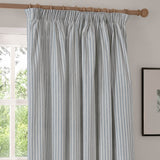 Hebden fully lined cotton pencil pleat curtain in mineral blue stripe, shown hung on a wooden pole beside a window in a living room setting.