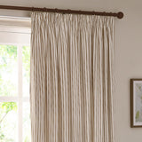 Hebden striped cotton pencil pleat curtains in natural beige and off-white, hung on a dark wooden pole beside a window in a living room.