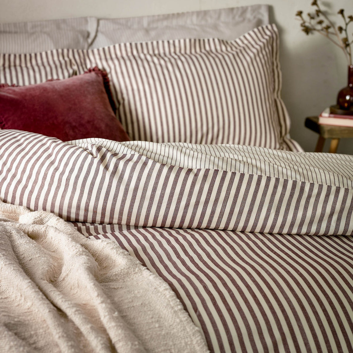 Hebden Cotton Melange Duvet Cover Set in Beetroot, showing cream and deep red striped bedding with matching pillowcases on a bed, styled with a cream throw and burgundy cushion.