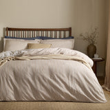 Hebden Cotton Melange Duvet Cover Set in caramel and cream stripe, styled on a bed with a wooden spindle headboard and navy pillowcases in a neutral bedroom.