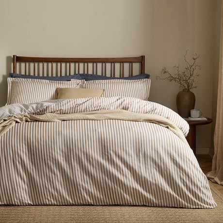 Hebden Cotton Melange Duvet Cover Set in caramel and cream stripe, styled on a bed with a wooden spindle headboard and navy pillowcases in a neutral bedroom.