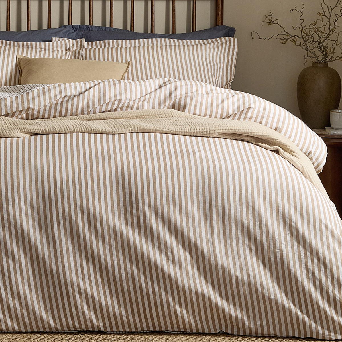 Hebden Cotton Melange Duvet Cover Set in Caramel, shown on a bed with striped cotton bedding, cream cushions and a woven throw in a bedroom setting.