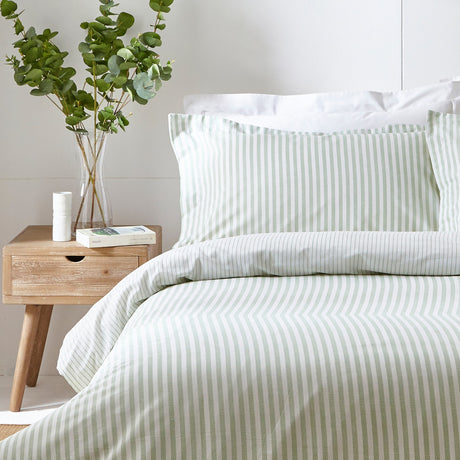Hebden Cotton Striped Duvet Cover Set in Eucalyptus, shown on a bed with matching pillowcases in a green and white woven stripe, styled in a bedroom beside a wooden nightstand and eucalyptus stems.