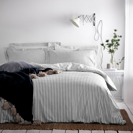 Hebden Cotton Striped Duvet Cover Set in grey and white on a bed with matching pillowcases, dark navy throw with tassels, in a bedroom with a wall lamp and side table.