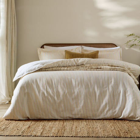 Hebden Cotton Melange Duvet Cover Set in honey yellow stripe on a bed with matching pillowcases, styled in a neutral bedroom with a woven rug and cane headboard.