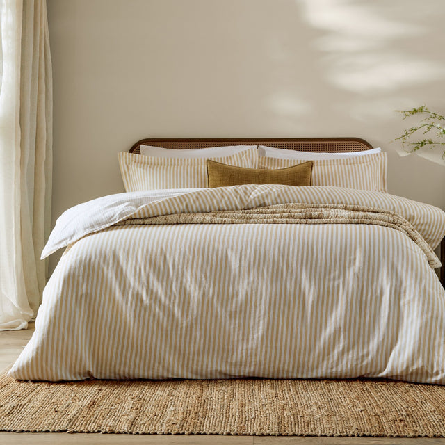 Hebden Cotton Melange Duvet Cover Set in honey yellow stripe on a bed with matching pillowcases, styled in a neutral bedroom with a woven rug and cane headboard.
