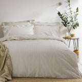 Hebden Cotton Striped Duvet Cover Set in natural beige and cream on a made bed with matching pillowcases, styled in a bedroom with a side table and potted plant.
