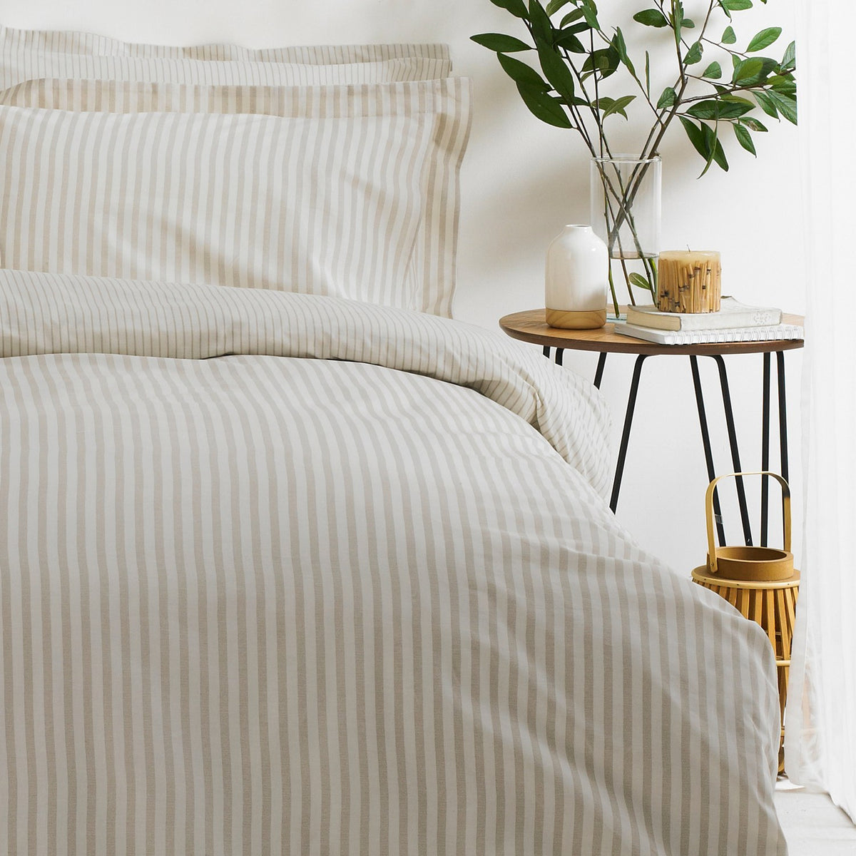 Hebden Cotton Striped Duvet Cover Set in natural beige and cream on a bed with matching pillowcase, styled in a bedroom beside a wooden side table with plant and candle.