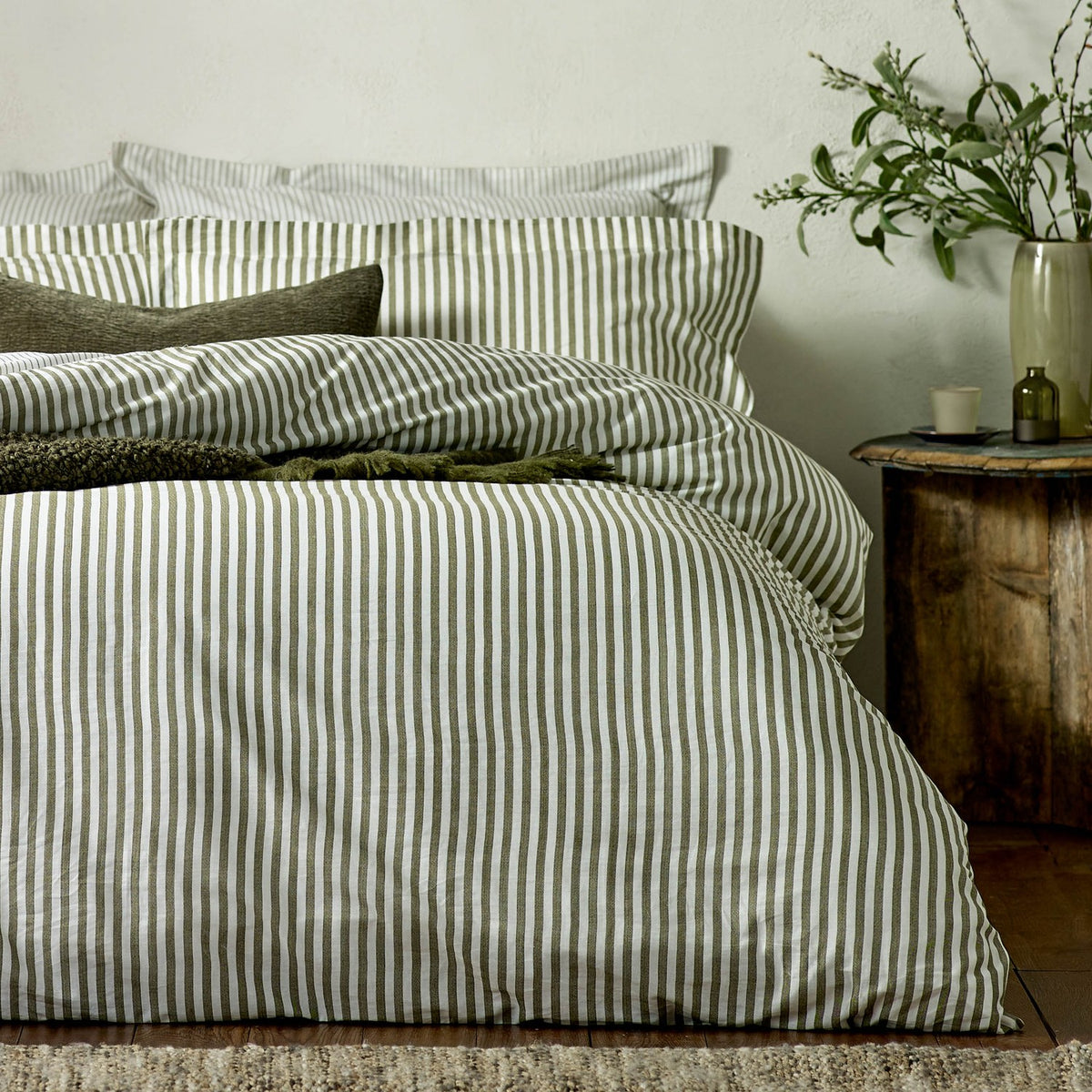 Hebden Cotton Melange Duvet Cover Set in olive green striped cotton, styled on a bed with matching pillowcases beside a wooden side table in a bedroom.