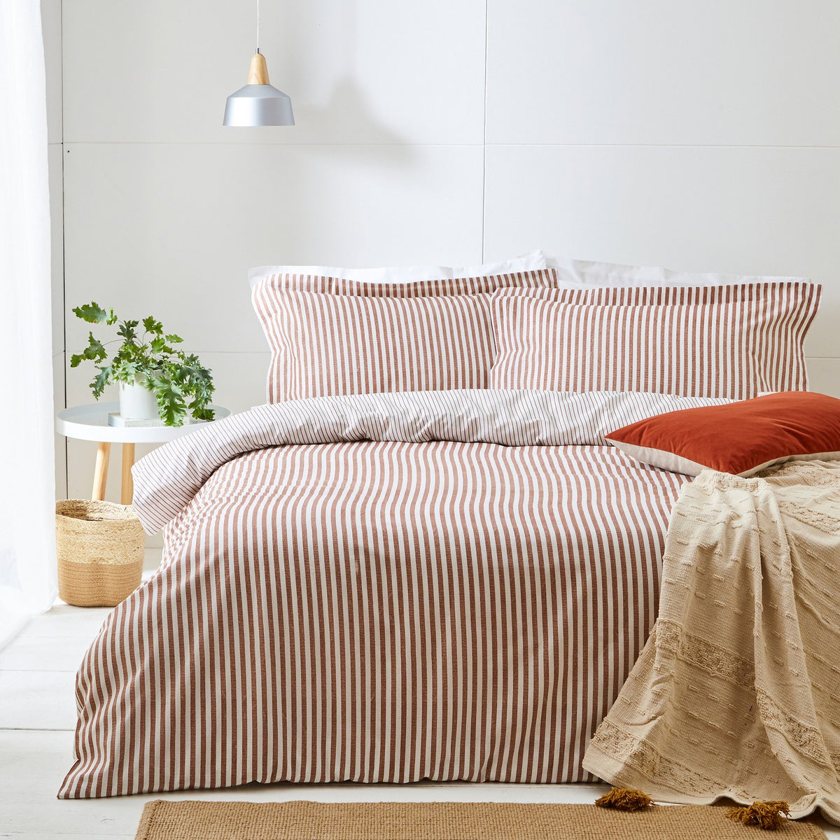Hebden Cotton Striped Duvet Cover Set in pecan on a bed with matching pillowcases, a rust cushion, and a beige textured throw in a light bedroom setting.