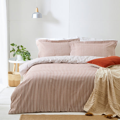Hebden Cotton Striped Duvet Cover Set in pecan on a bed with matching pillowcases, a rust cushion, and a beige textured throw in a light bedroom setting.