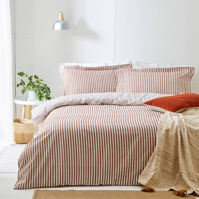 Hebden Cotton Striped Duvet Cover Set in pecan on a bed with matching pillowcases, a rust cushion, and a beige textured throw in a light bedroom setting.
