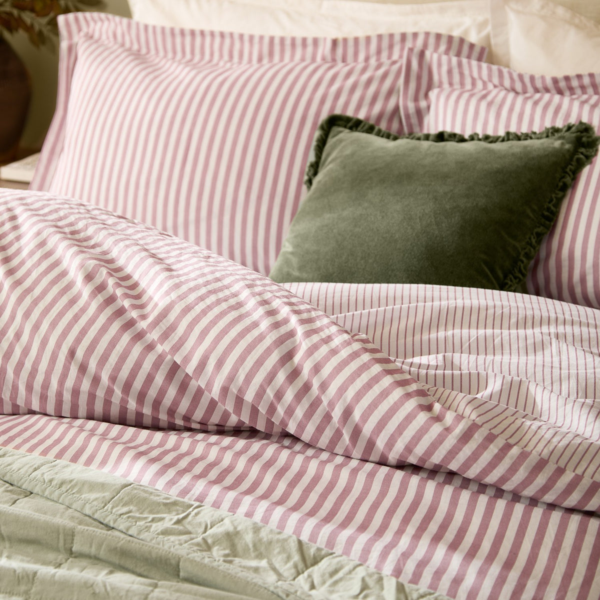 Hebden Cotton Melange Duvet Cover Set in Rosewood on a bed with matching pink and white striped pillowcases, sage green quilted throw, and a green velvet cushion.