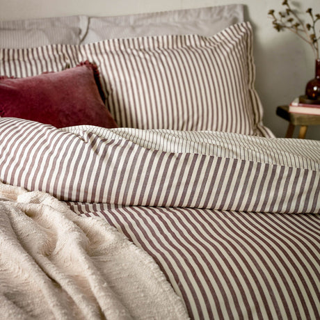 Hebden Striped Cotton Pillowcase Pair in Beetroot, shown styled on a bedroom bed with matching striped duvet cover, red velvet cushion, and cream throw.