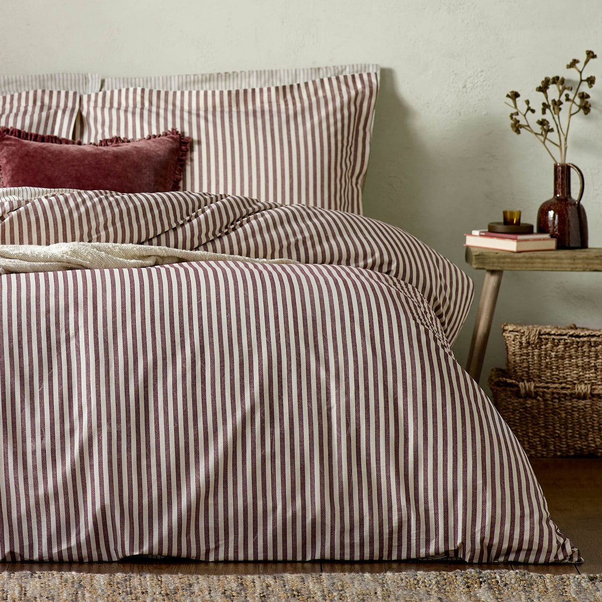 Bedroom scene with Hebden striped cotton pillowcases in beetroot red and off-white, dressing a bed with matching striped duvet cover and a red velvet cushion.