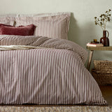 Bedroom scene with Hebden striped cotton pillowcases in beetroot red and off-white, dressing a bed with matching striped duvet cover and a red velvet cushion.