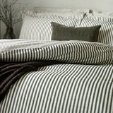 Hebden Striped Cotton Pillowcase Pair in black on a bed, layered with matching striped shams, a plain charcoal cushion and a chunky knit throw in a bedroom setting.