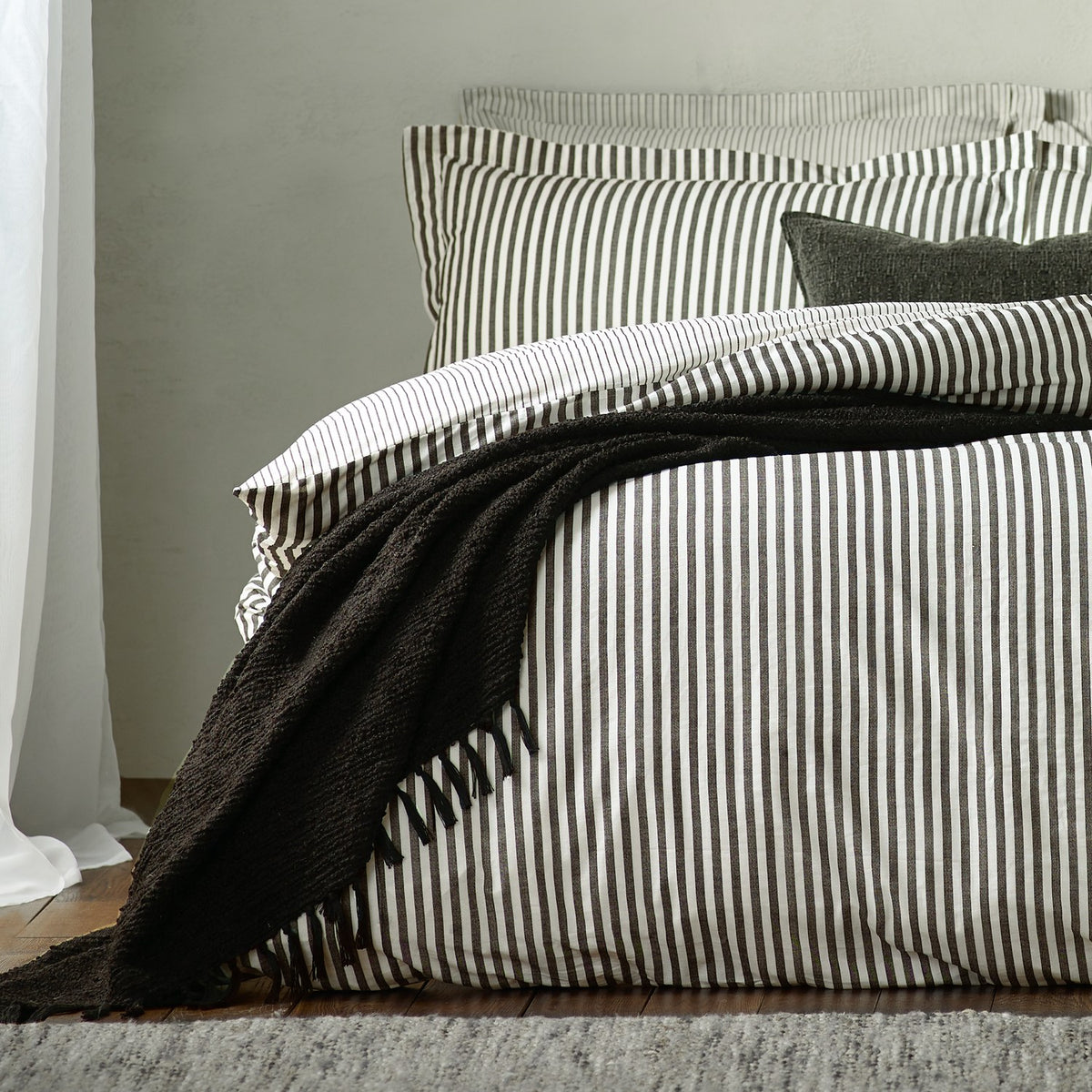 Hebden Striped Cotton Pillowcase Pair in Black, shown styled on a bed with matching striped duvet cover and a textured black throw in a bedroom setting.