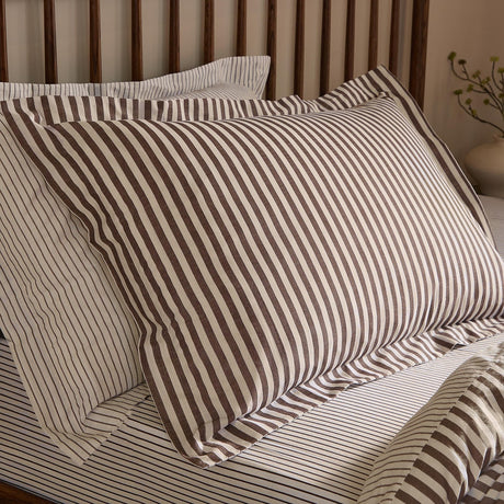 Hebden Reversible Cotton Pillowcase Pair in Espresso Brown, striped brown and cream cotton pillow shown on a bed with a wooden slatted headboard in a bedroom setting.