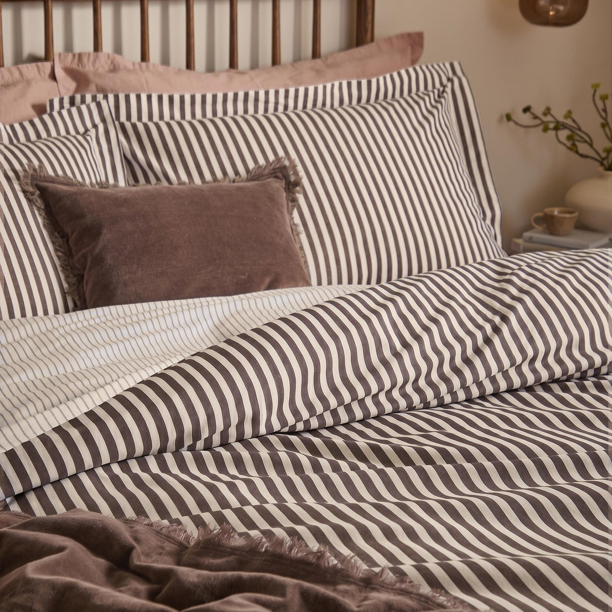 Hebden Reversible Cotton Pillowcase Pair in espresso brown and cream stripe, styled on a bed with a velvet cushion and brown throw in a bedroom setting.
