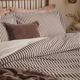 Hebden Reversible Cotton Pillowcase Pair in espresso brown and cream stripe, styled on a bed with a velvet cushion and brown throw in a bedroom setting.