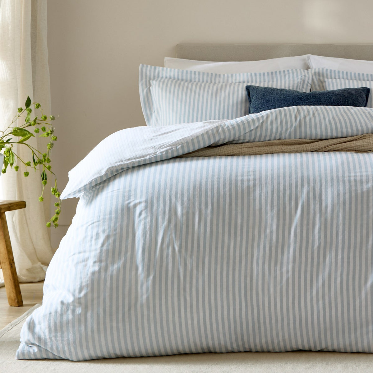 Bed styled with Hebden Melange Cotton Pillowcase Pair in Mineral Blue, soft-blue striped pillowcases and duvet cover, and navy accent cushions in a neutral bedroom.