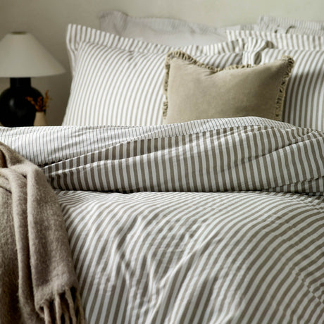 Hebden Striped Cotton Pillowcase Pair in mole-brown and white on a bed, styled with matching pinstripe bedding, a textured cushion and a knitted throw.