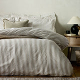 Bed dressed in Hebden Striped Cotton Pillowcase Pair in mole-brown and white stripes, styled with a matching duvet cover, textured cushion, and wooden bedside table with lamp.